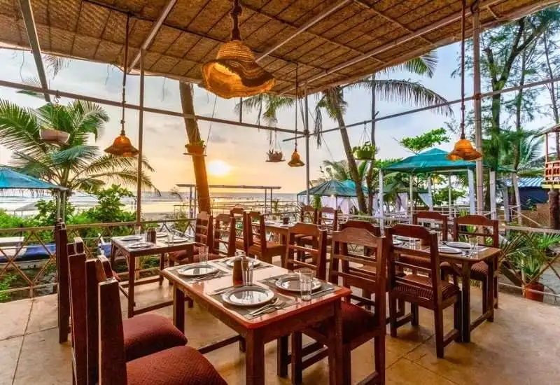 Open-air beachside dining area with wooden tables, palm trees, and sunset view at the best resort in Morjim